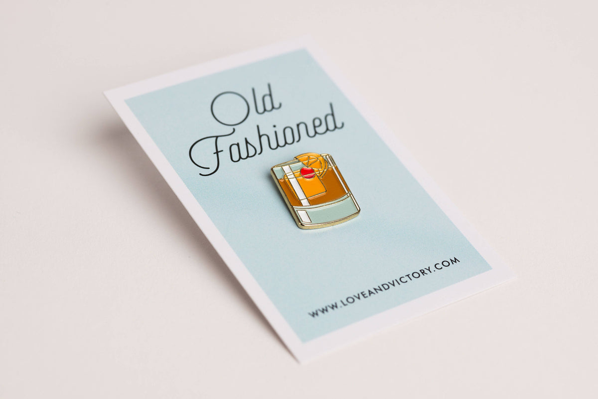 Enamel pin of an old-fashioned cocktail on a card with &#39;Old Fashioned&#39; text and &#39;Love and Victory&#39; website.