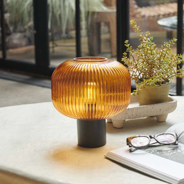 Decorative table lamp with ribbed design on a surface with glasses and a book in the background.