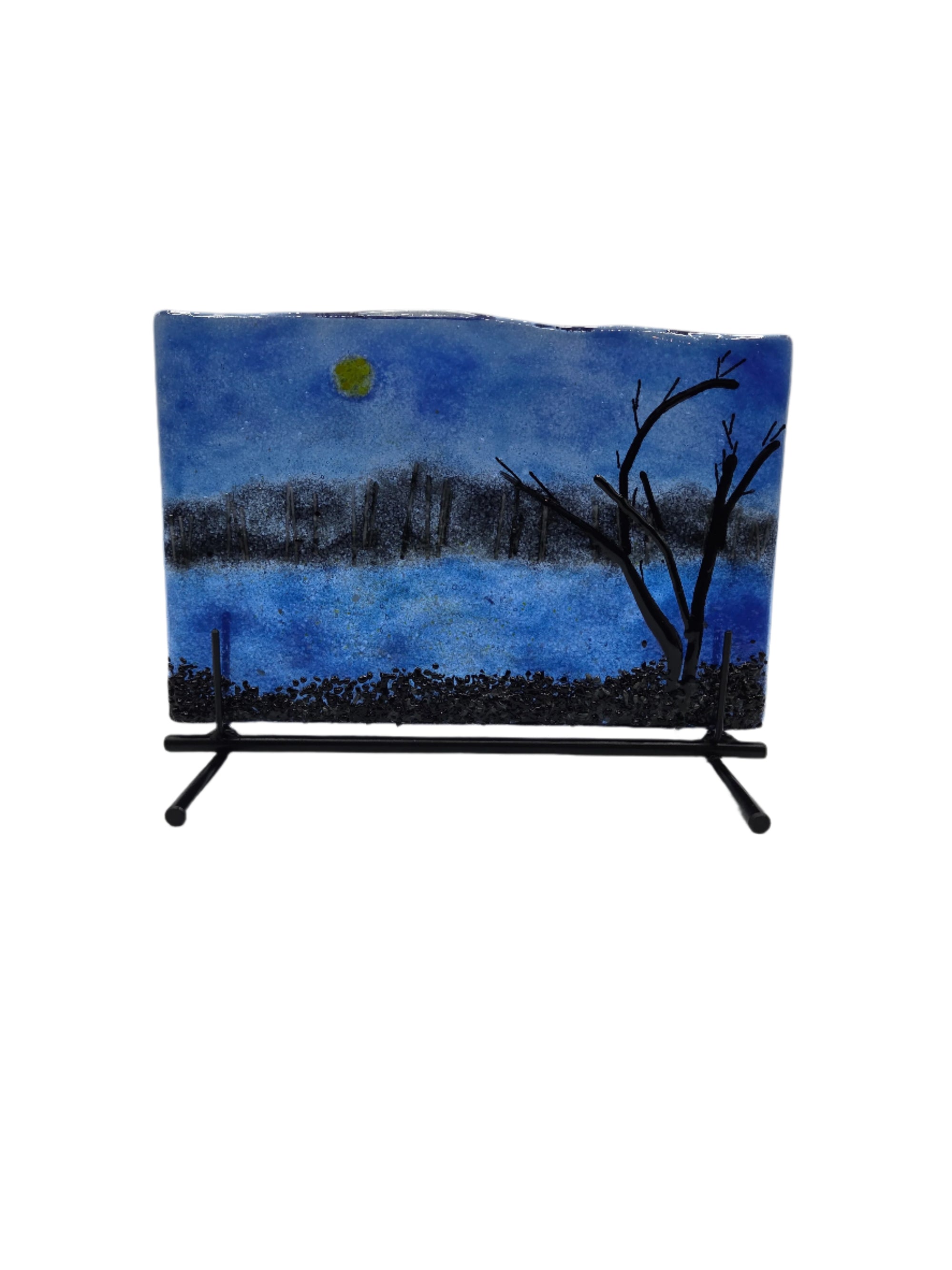 Fused glass art piece with trees and a moon on a blue background on a stand.