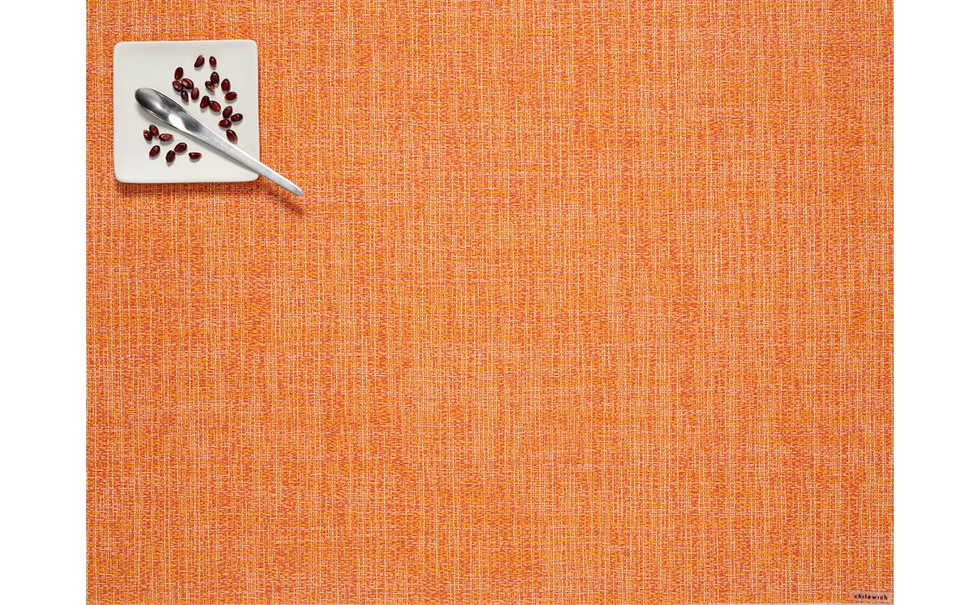 Orange placemat with a square coaster featuring a spoon and pomegranate seeds on a white background