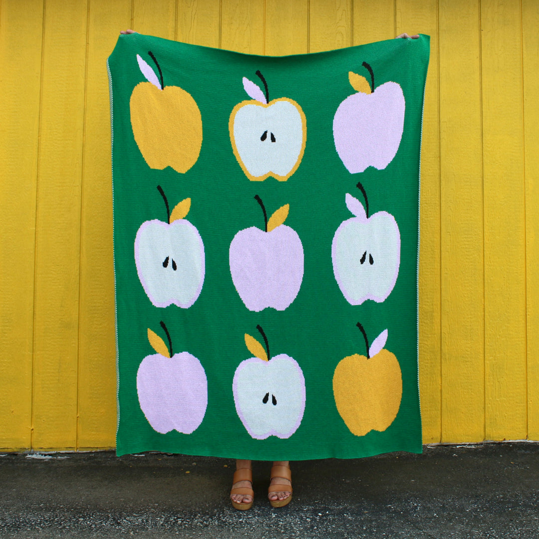 Person holding a green blanket with colorful apple pattern against a yellow wall.