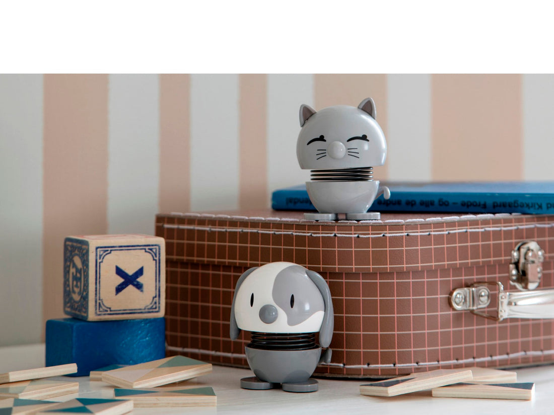 A gray cat-shaped figurine and a gray dog figurine on a striped wall background with children&#39;s toys around.