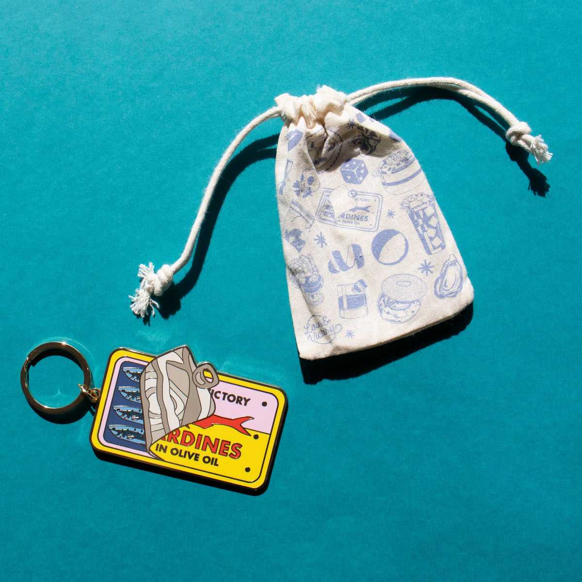 Keychain with vintage sardines label design and small drawstring bag on a teal background