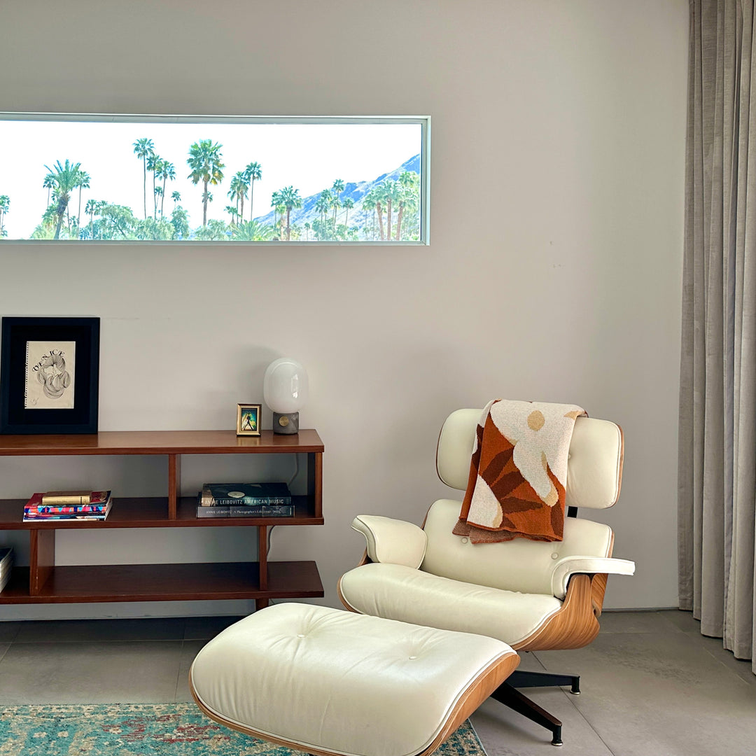 Modern living room with a white armchair with an orange and cream large floral pattern hanging over the back. An ottoman, a wooden side table, and wall art.