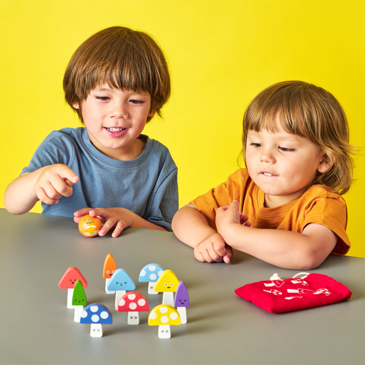 Two children playing with colorful toy mushrooms on a gray table against a yellow background