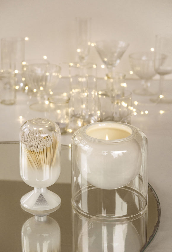 White candle in a glass holder with matchbox on a reflective surface with blurred lights in the background