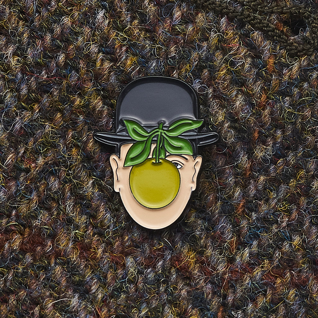 Pin with a design of a face wearing a bowler hat with green leaves and a yellow fruit on a textured brown background