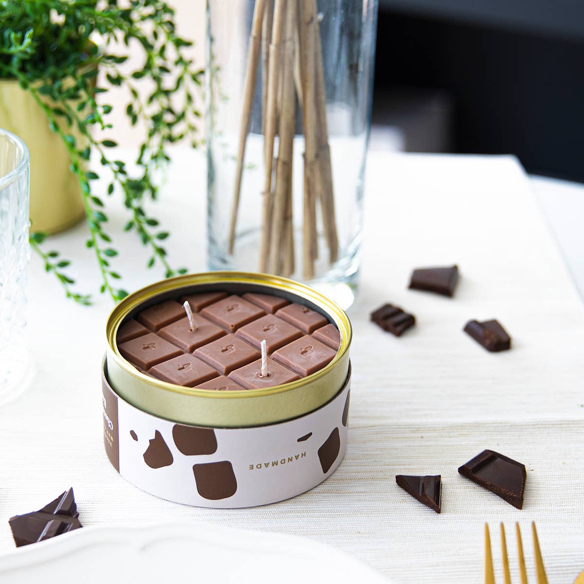 Chocolate-scented candles in a tin on a table with chocolate pieces and a plant in the background.