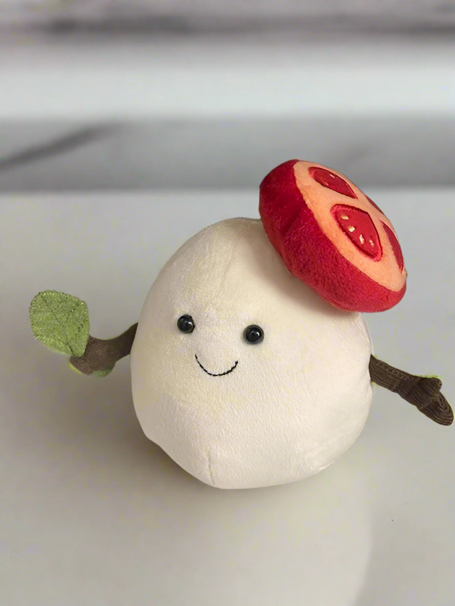 Plush toy resembling a white ball of mozzarella with a red tomato on top, black eyes, and a green leaf on a light gray background.